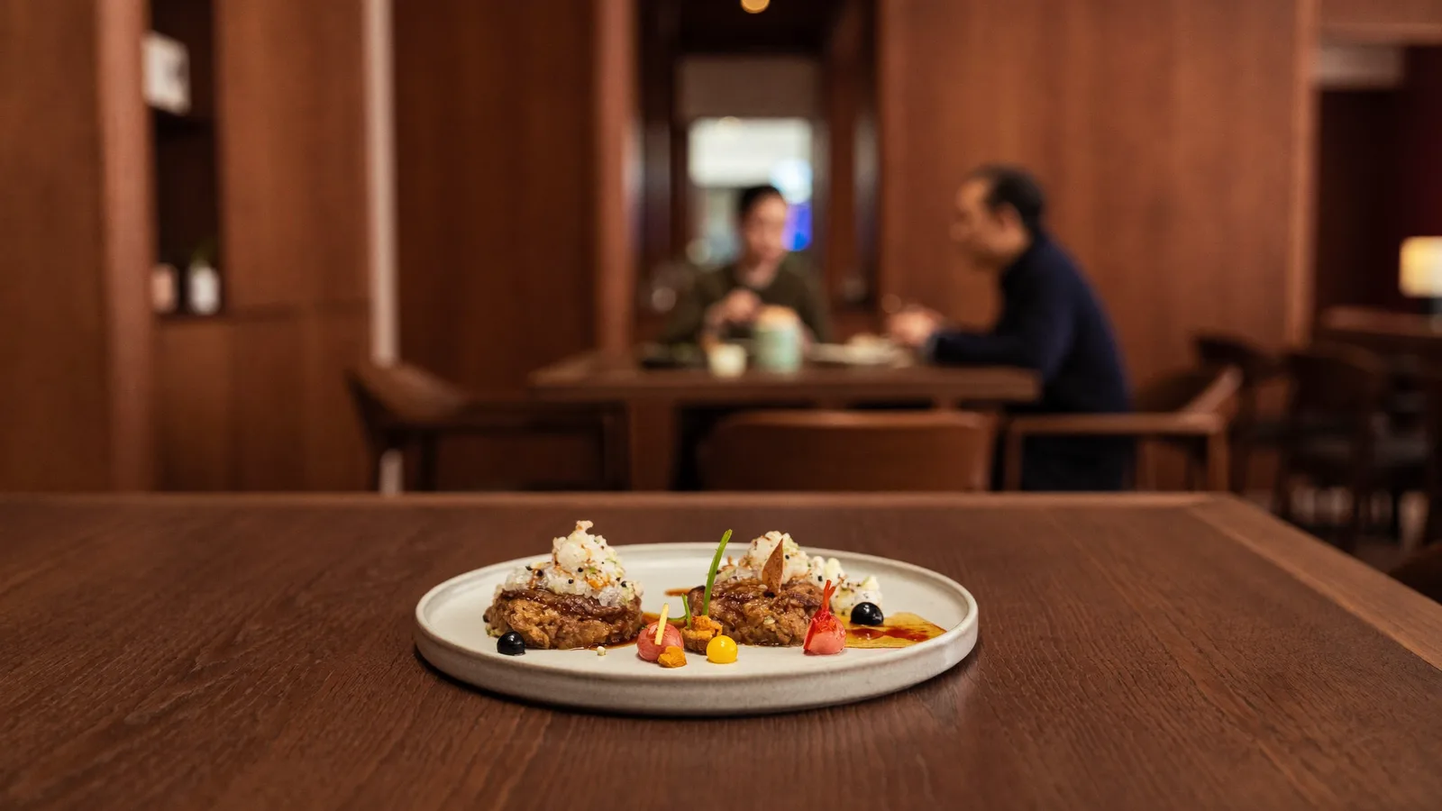 Illustrative image of a fine-dining tasting menu course, an elegant French-Japanese plated dish on a rich walnut table in a warmly lit modern restaurant interior, a local Hong Kong Chinese diner softly blurred in the background, not a photo of the actual restaurant or dishes