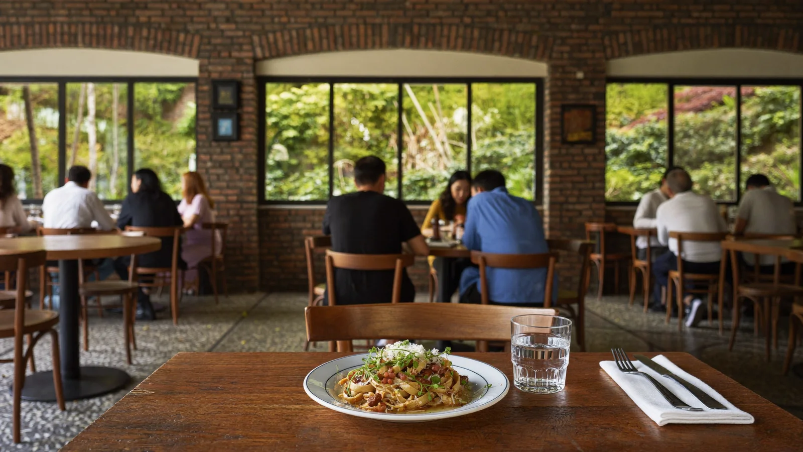 Illustrative image of a cosy cottage-style bistro dining room with exposed brick walls and garden views through large windows, a plated Western dish on a wooden table in the foreground, local Hong Kong Chinese diners softly blurred in the background, illustrative image for this Mei Foo bistro guide, not a photo of the actual venue