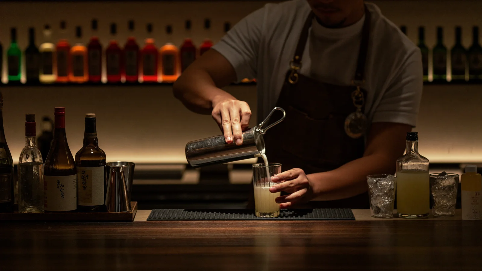 A moody, minimalist Hong Kong cocktail bar interior with unlabelled bottles and a bartender crafting a drink at a dark wooden counter, warm low lighting, illustrative image for this Bar Mind Chapter III cocktail menu guide, not a photo of the actual venue