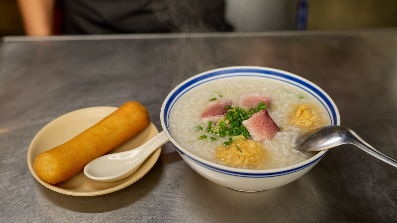 A steaming bowl of Cantonese congee with spring onion and a fried dough stick on a stainless steel table - illustrative hero for this Hong Kong congee guide, not a photo of a specific shop