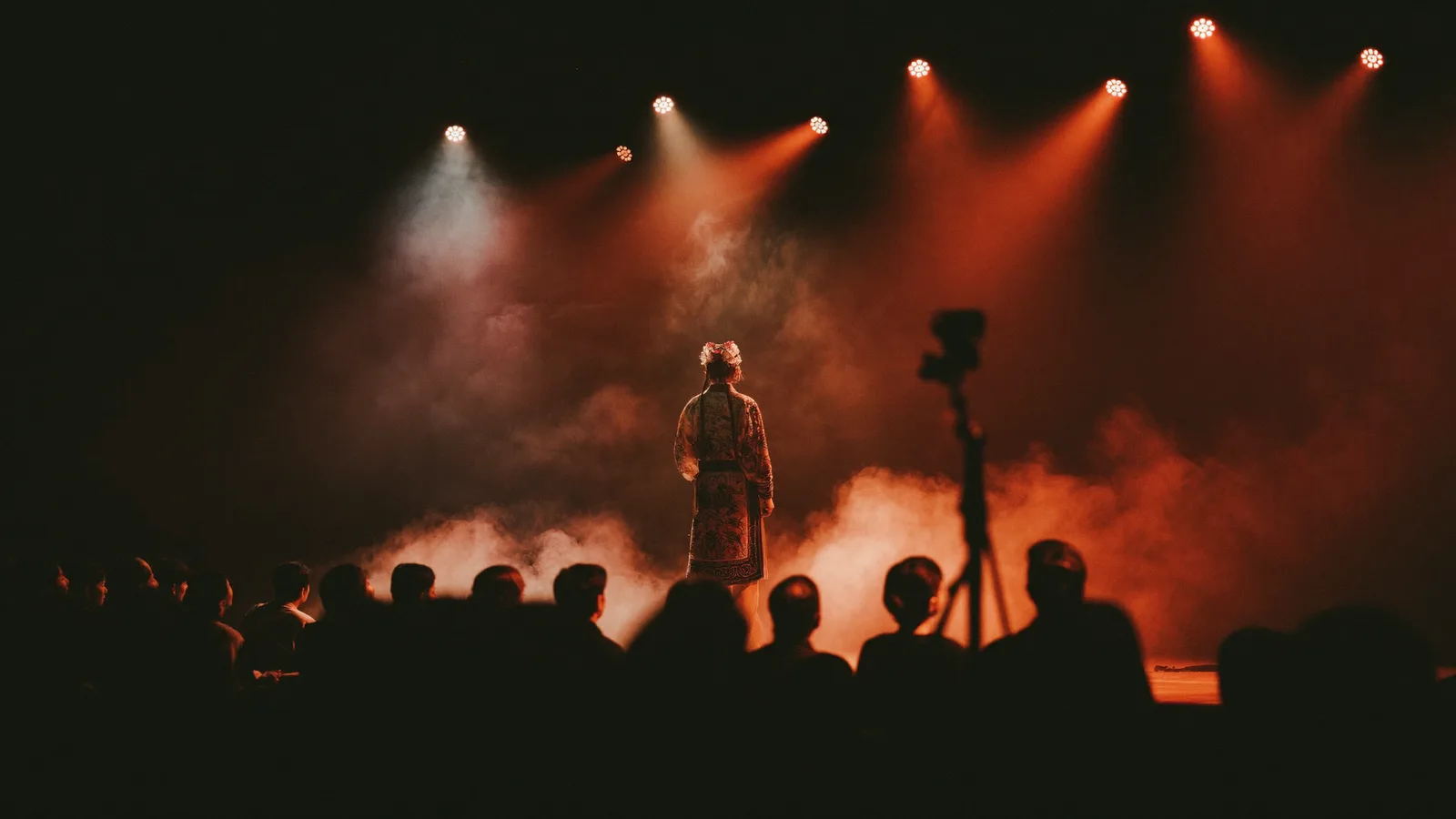 Illustrative image of a Chinese opera stage performance, with an ornately costumed performer in soft focus under warm stage lighting inside a black-box theatre, a local Hong Kong Chinese audience blurred in the darkened seats, not a photo of the actual performers or venue