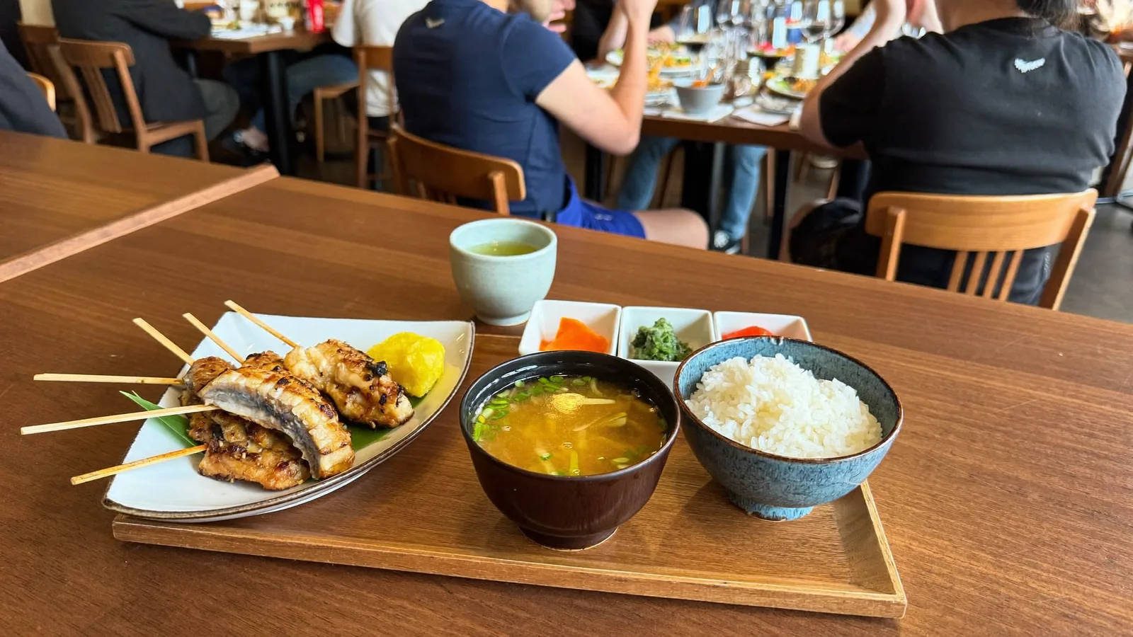 A Japanese-style teishoku set tray with grilled fish, miso soup, pickles and rice on a wooden table, local Hong Kong Chinese diners in the background, illustrative image for this CHIU San Kennedy Town restaurant guide, not a photo of the actual restaurant or dishes