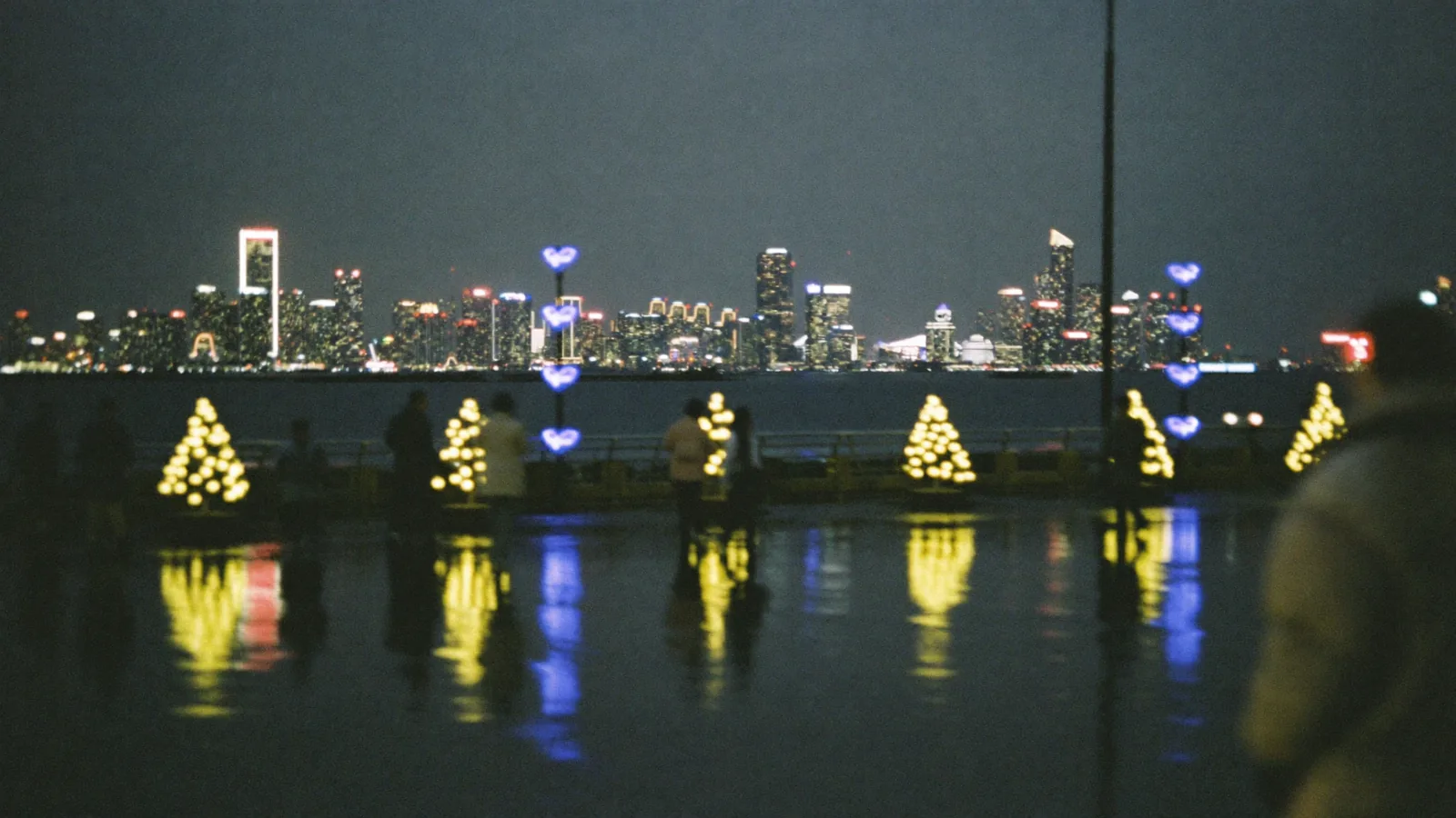 香港海濱夜晚，小型聖誕樹亮住暖色燈串，行人剪影走過，此為聖誕指南示意圖，並非某指定場地實照