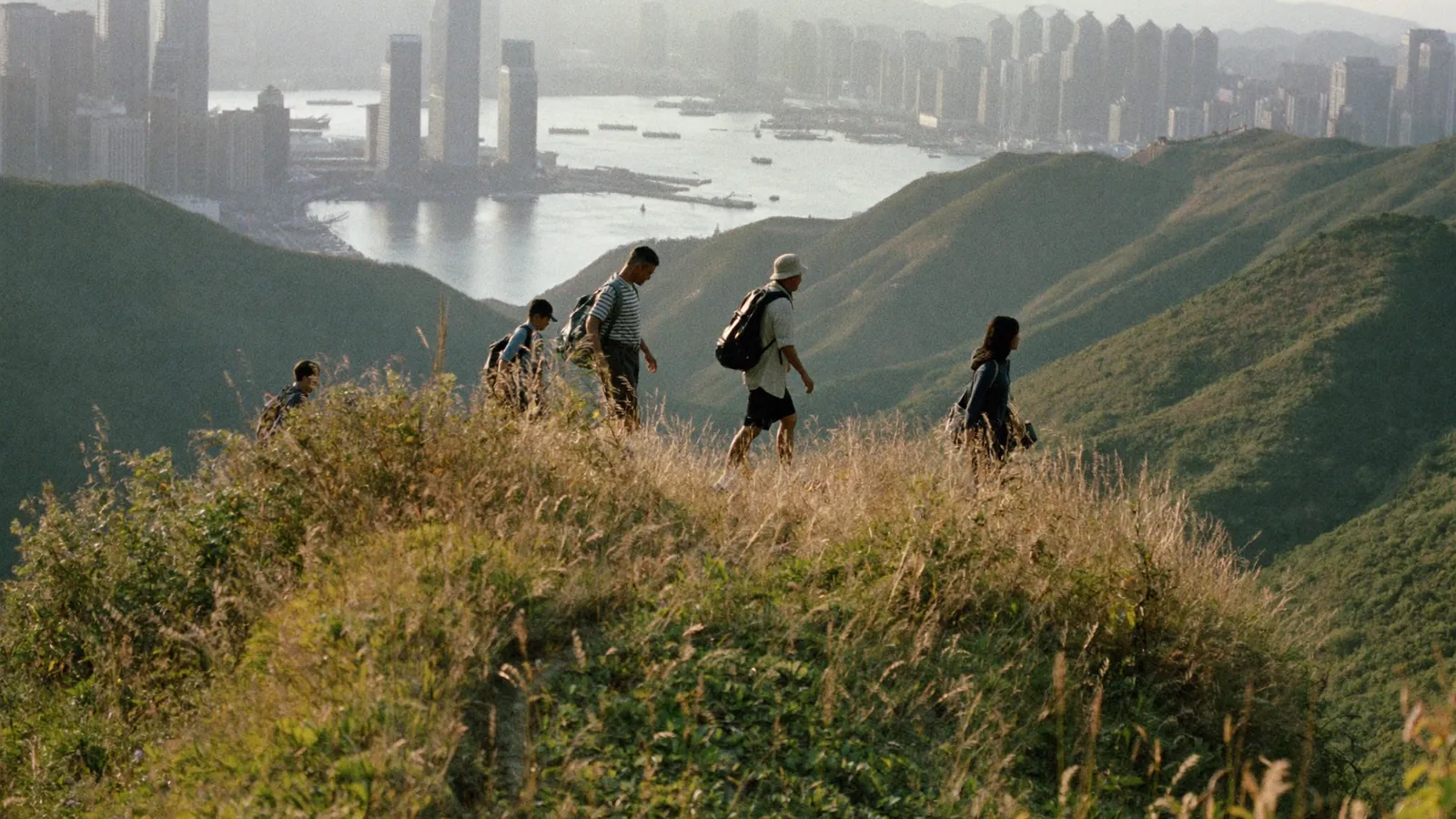 A small group of hikers walking a grassy ridge path high above a hazy Hong Kong harbour in late afternoon light - an illustrative image for this Chung Yeung Festival long-weekend guide, not a photograph of a specific trail