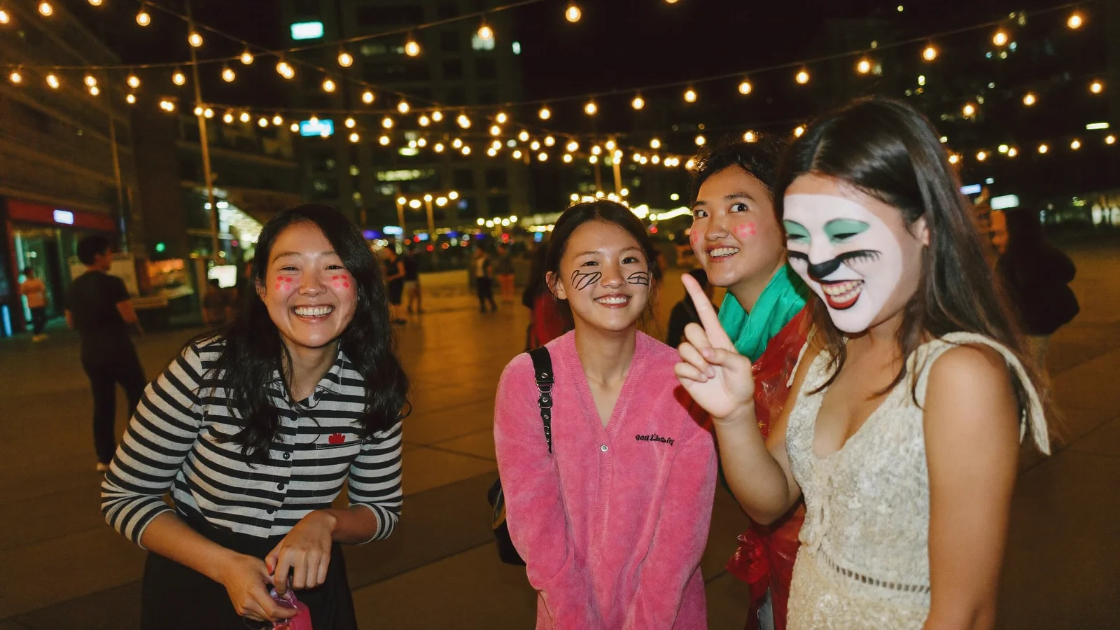 A small group of local Hong Kong Chinese friends laughing together at night with playful Halloween face paint and costumes, standing in an open plaza with warm string lights and a softly blurred city backdrop - illustrative hero for this Hong Kong citywide Halloween guide, not a photo of a past event