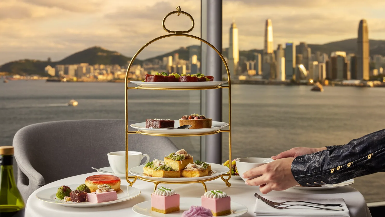 An elegant gold-and-white afternoon tea stand with delicate Italian savoury bites and pastel-coloured desserts, set at a harbour-view hotel restaurant table with Victoria Harbour visible through the window and a local Hong Kong Chinese couple enjoying tea, illustrative image for this Cucina afternoon tea guide, not a photo of the actual dishes