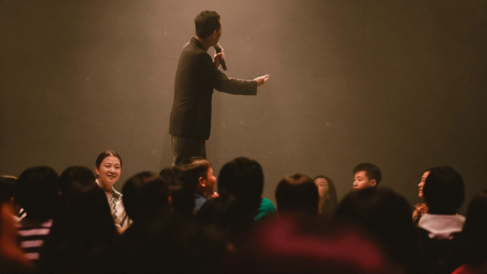 A stand-up comedian silhouetted on a small stage under a single spotlight with a microphone stand, local Hong Kong Chinese audience seated in rows in the foreground, illustrative image for this Des Bishop comedy guide, not a photo of the actual show or performer