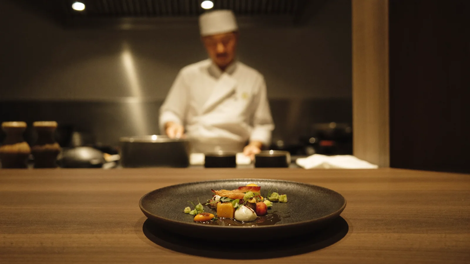 An elegantly plated tasting-menu course on a dark ceramic dish in a warm, minimalist fine-dining room with an open kitchen in the background, illustrative image for this Ébauche restaurant guide, not a photo of the actual venue