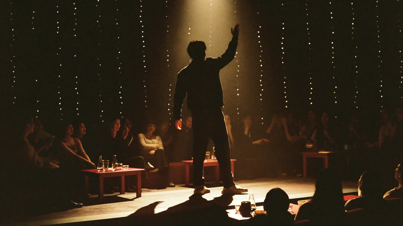 An intimate comedy club stage with a silhouetted performer mid-gesture under a warm spotlight, a small audience of local Hong Kong Chinese people laughing and enjoying the show in the foreground, illustrative image for this Elena Gabrielle comedy show guide at BaseHall 2, not a photo of the actual performer or venue