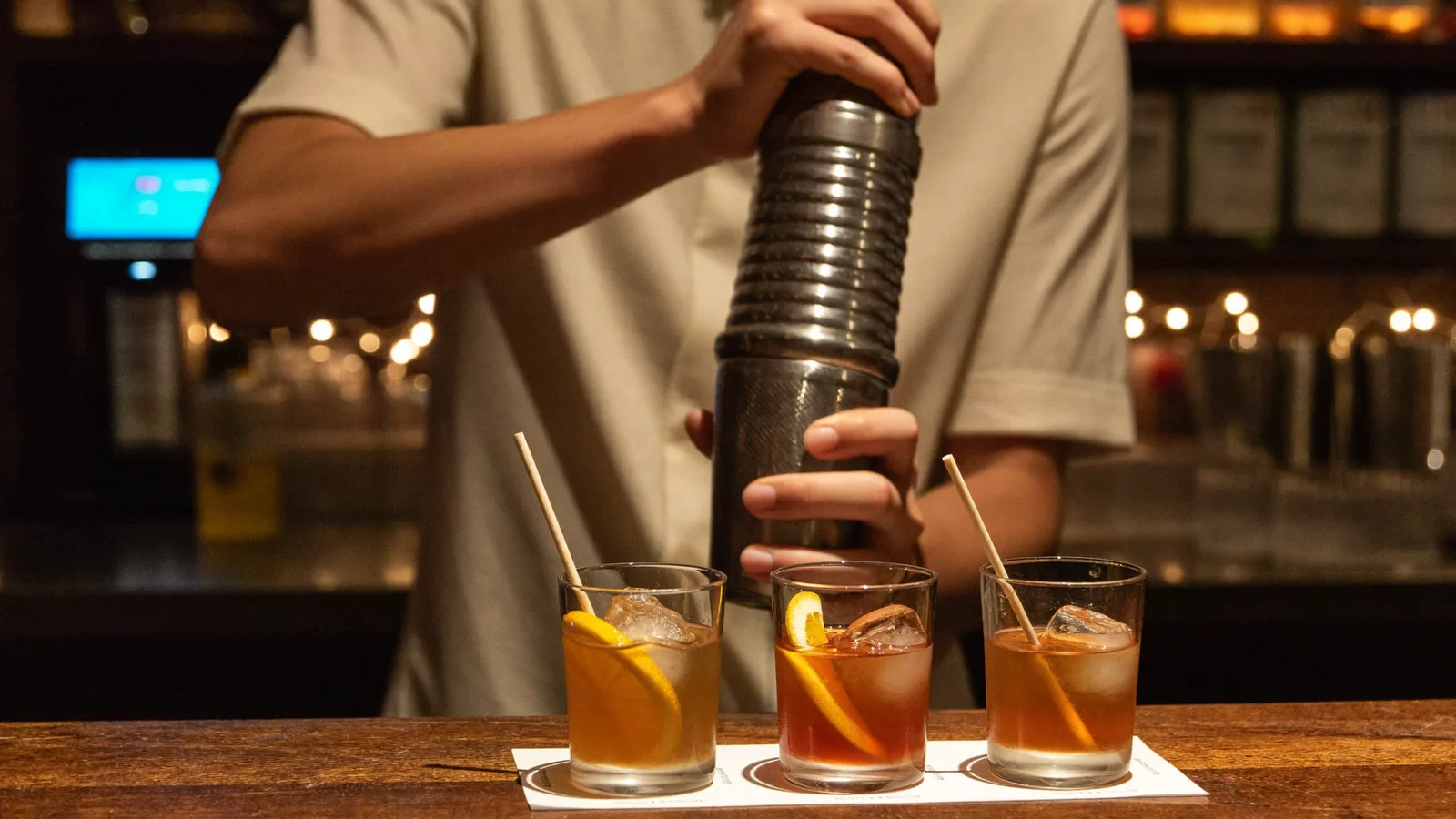 A bartender shaking a cocktail behind a warm, backlit bar counter with glassware and citrus garnishes, illustrating a guide to the new Central cocktail bar Fello - an illustrative image, not a photo of the actual bar