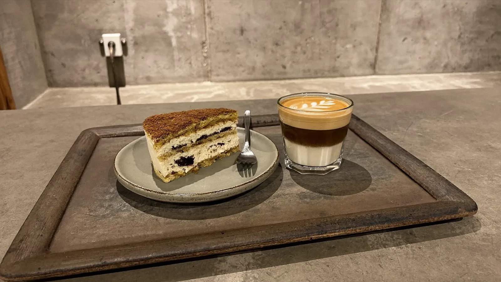 A slice of tiramisu cake and a layered latte with leaf-pattern latte art on a wooden tray in an industrial-chic concrete cafe interior, illustrative image for this Gettin Kennedy Town opening guide, not a photo of the actual venue or drinks