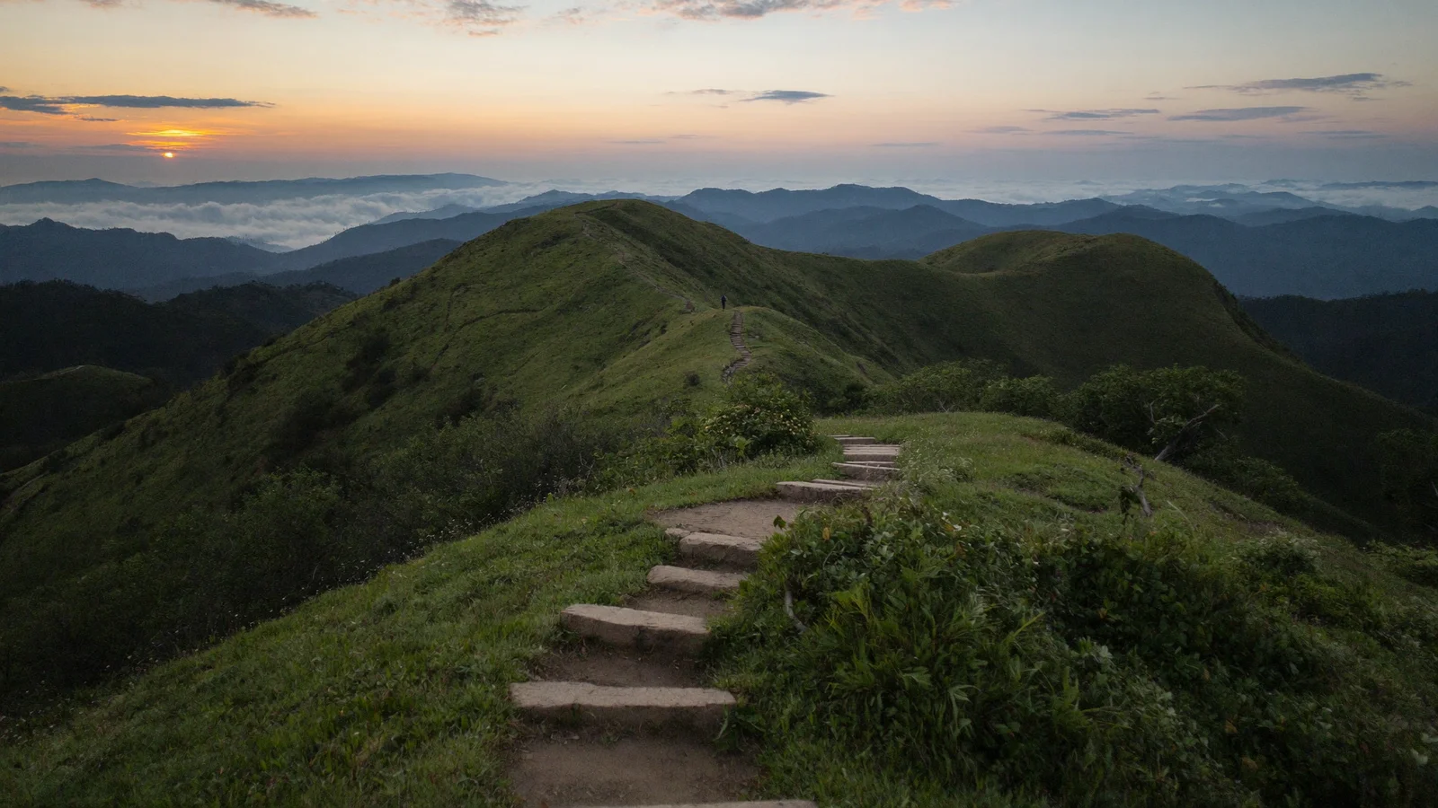 A rugged mountain trail on Lantau Island Hong Kong at dawn, steep stone steps winding up a green hillside with misty peaks in the distance, a single distant trail runner silhouette on the path, illustrative image for this Hilly Grail trail race guide, not a photo of the actual event