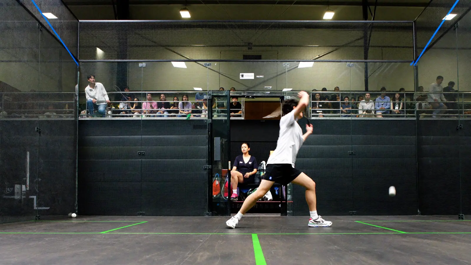 A squash player mid-swing inside a glass-panelled show court with spectators watching from a stand above, local Hong Kong Chinese players and audience, illustrative image for this Hong Kong Squash Cup guide, not a photo of the actual tournament or players
