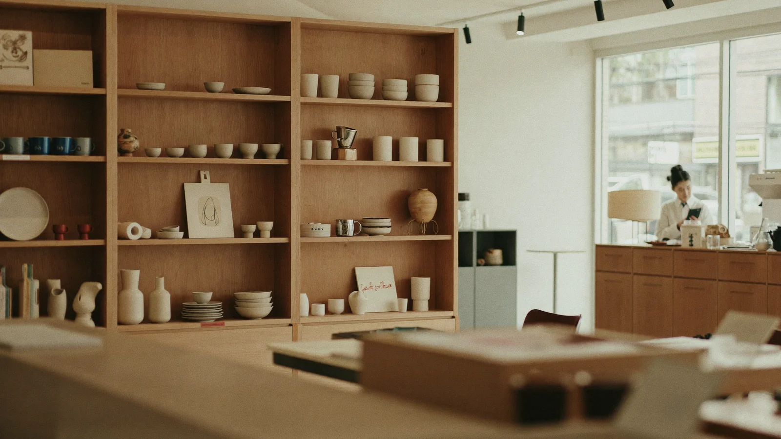 A minimalist Japanese-curated homeware and furniture store interior with warm wooden shelving, ceramics and design objects neatly displayed, a small cafe counter visible at the back with a local Hong Kong Chinese barista softly blurred, large street-facing windows letting in natural light, illustrative image for this Kwun Tong shop guide, not a photo of the actual venue