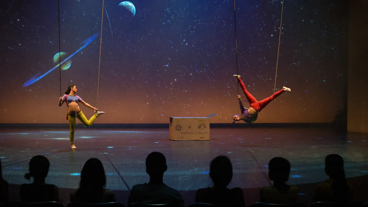 Aerial dancers soaring above a theatre stage lit with cosmic star projections as families watch the International Arts Carnival in Hong Kong