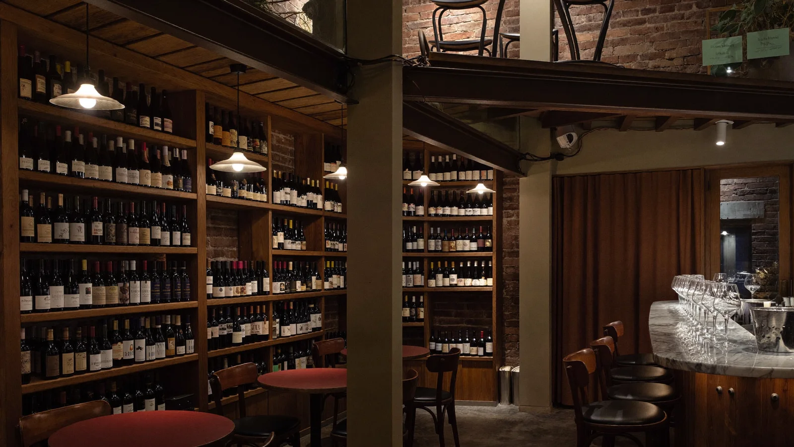 A cosy, warmly lit two-storey wine bar interior with wooden shelving of wine bottles along the walls, a marble bar counter with wine glasses catching warm light, illustrative image for this James Suckling Wine Central guide, not a photo of the actual venue