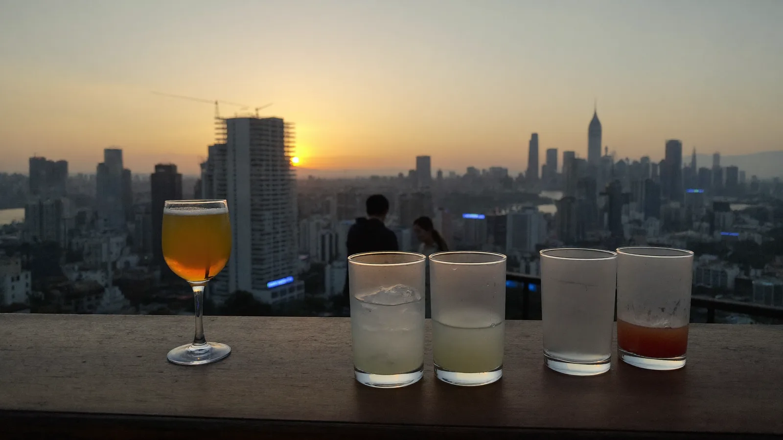 A rooftop bar at sunset with colourful cocktails on the counter and a city skyline glowing in the background, illustrative hero for this Kai Tak rooftop bar guide, not a photo of the actual venue