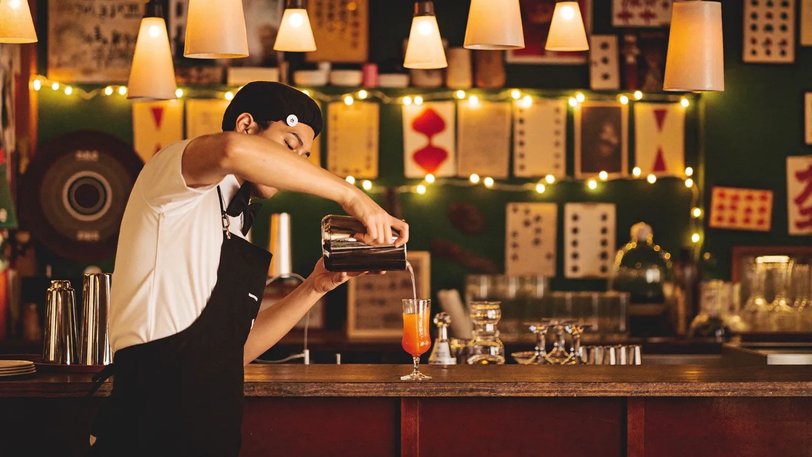 A local Hong Kong Chinese bartender pouring a colourful cocktail at a retro-themed bar counter lined with playing cards and warm string lights, illustrative image for this Kidult Tsim Sha Tsui bar guide, not a photo of the actual venue