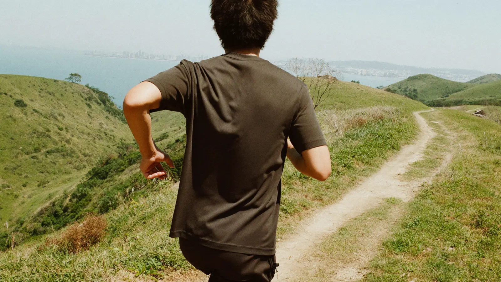 A local Hong Kong Chinese trail runner shown from behind running along a mountain ridge trail with green outlying-island peaks and sea haze in the distance, illustrative image for this Lantau 2 Peaks trail race guide, not a photo of the actual event or any real participant
