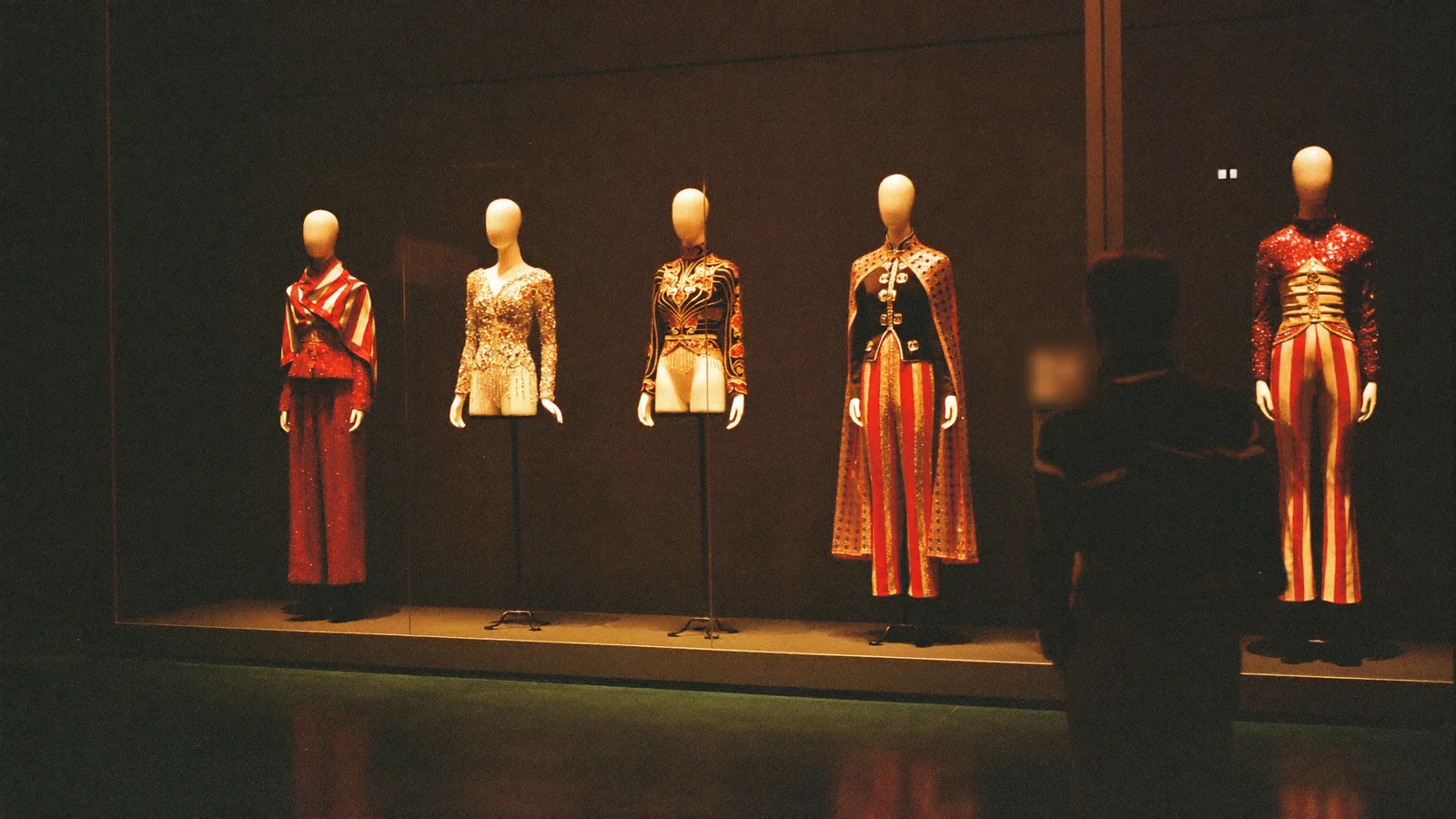 Glittering 1990s Cantopop stage costumes on faceless display mannequins under warm spotlights in a darkened museum gallery, with a single visitor silhouetted in front of them - an illustrative image for this guide to the Leslie Cheung exhibition, not a photograph of the Hong Kong Heritage Museum galleries