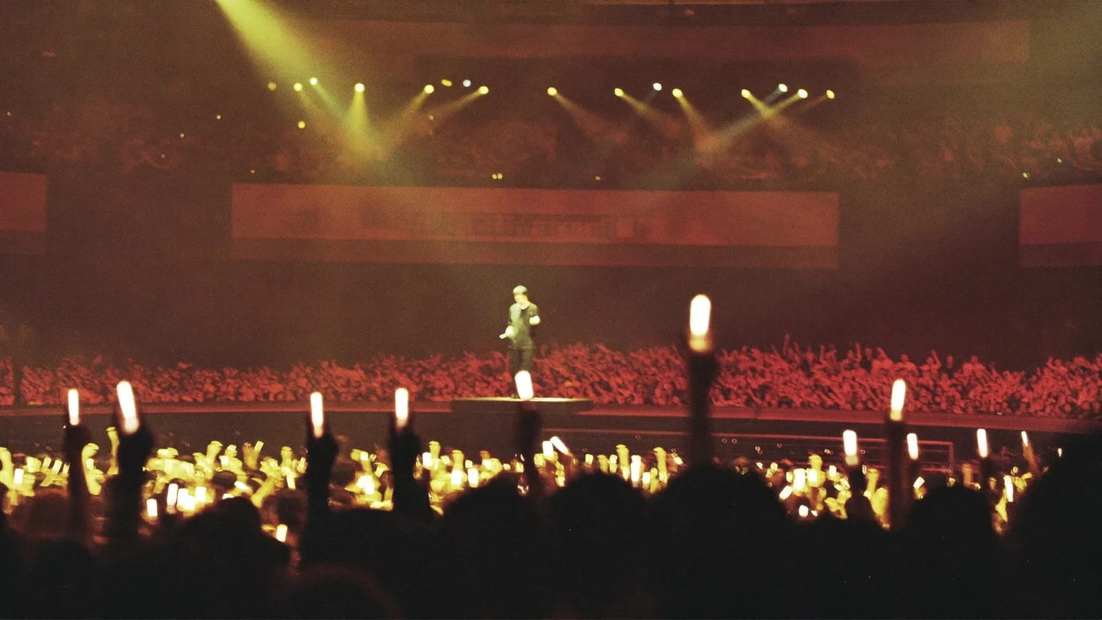 A packed indoor arena concert stage lit in warm gold and red with a single spotlit performer - an illustrative hero image for this Louis Koo Hong Kong Coliseum guide, not a photo of the actual show