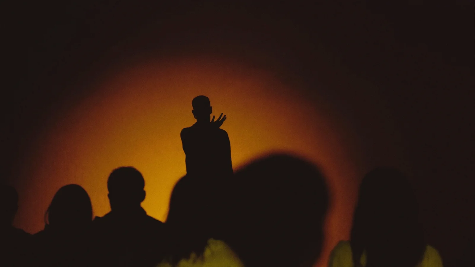 An intimate late-night comedy stage lit by a single warm amber spotlight, a faceless silhouetted stand-up comedian mid-gesture, blurred silhouettes of an audience's heads in the foreground, illustrative image for this Mohammed Magdi comedy show guide at Fa Gai, not a photo of the actual performer or venue
