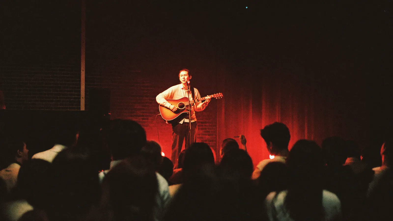 A small live music venue stage with warm stage lighting and a solo musician performing with a guitar, local Hong Kong Chinese audience watching, illustrative image for this mui zyu at The Wanch concert guide, not a photo of the actual performer or venue