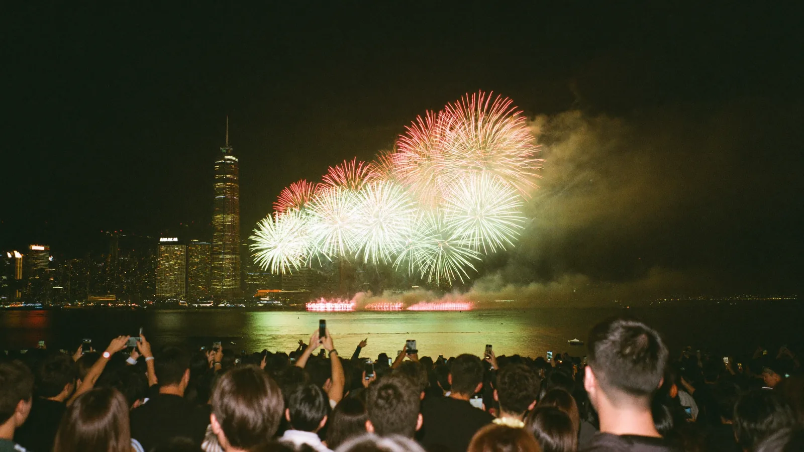 香港海濱長廊上嘅人群望住維港上空金色同紅色嘅煙花，此為國慶煙花指南嘅示意圖片，並非2026年匯演實景