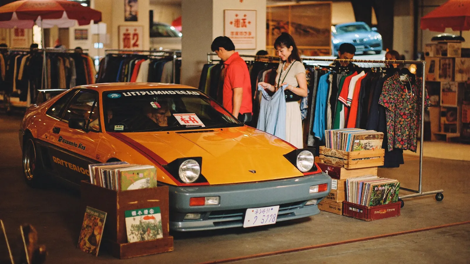 一個充滿活力嘅復古節慶場館，一部90年代日本經典跑車喺暖色調展燈下展出，背景係模糊嘅Y2K色彩繽紛古著同一箱箱黑膠唱片，一對本地香港華人情侶一齊行街睇貨，此為南豐紗廠RE-LOVE Festival Retro Rewind指南嘅示意圖片，並非實際活動相片