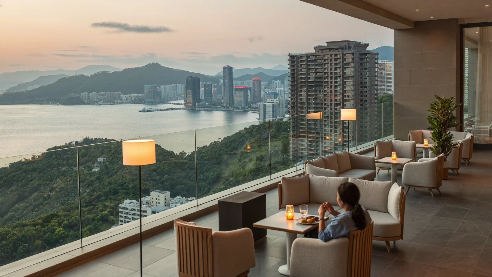 A stylish hotel rooftop terrace at dusk overlooking a bay ringed by green hills, illustrating a guide to The Royal Garden Kowloon East - an illustrative image, not a photo of the actual hotel