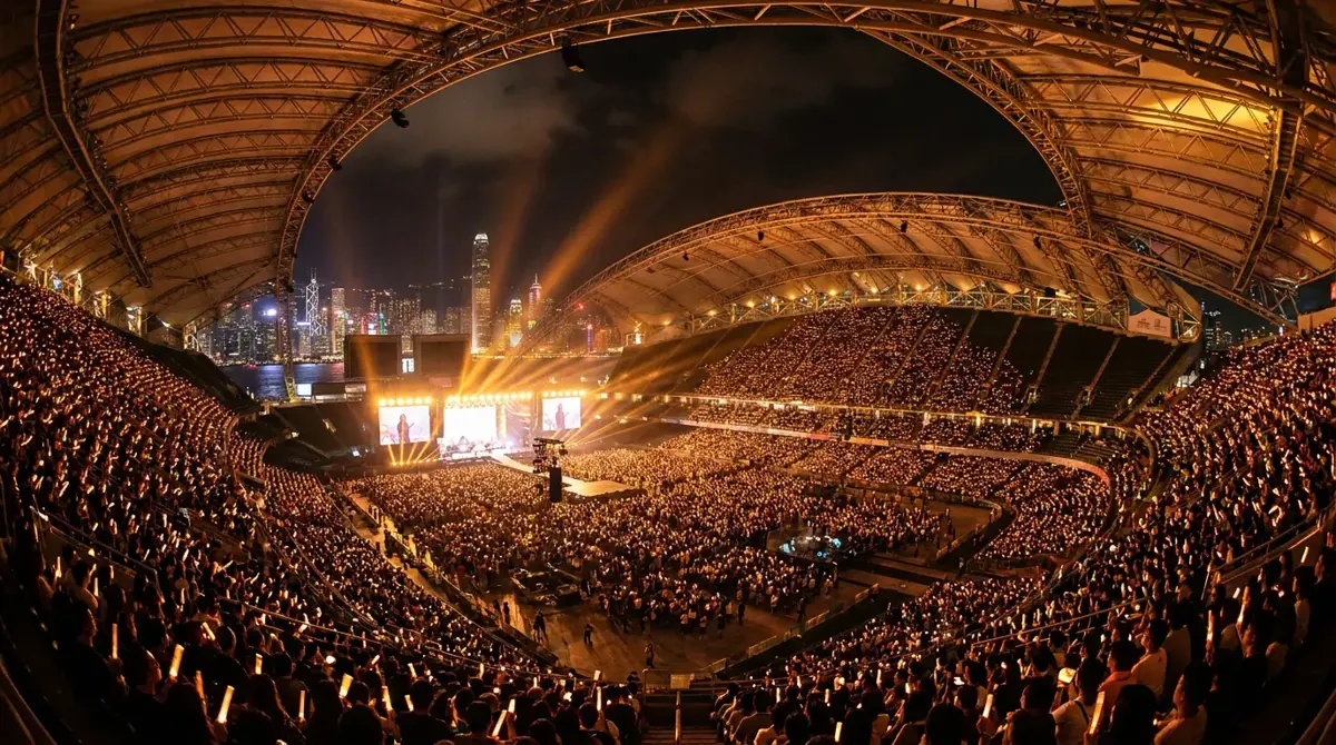 A packed 50,000-seat stadium at night in Hong Kong during a major Cantopop concert, with amber stage lighting, a glowing stage and the Kowloon skyline beyond