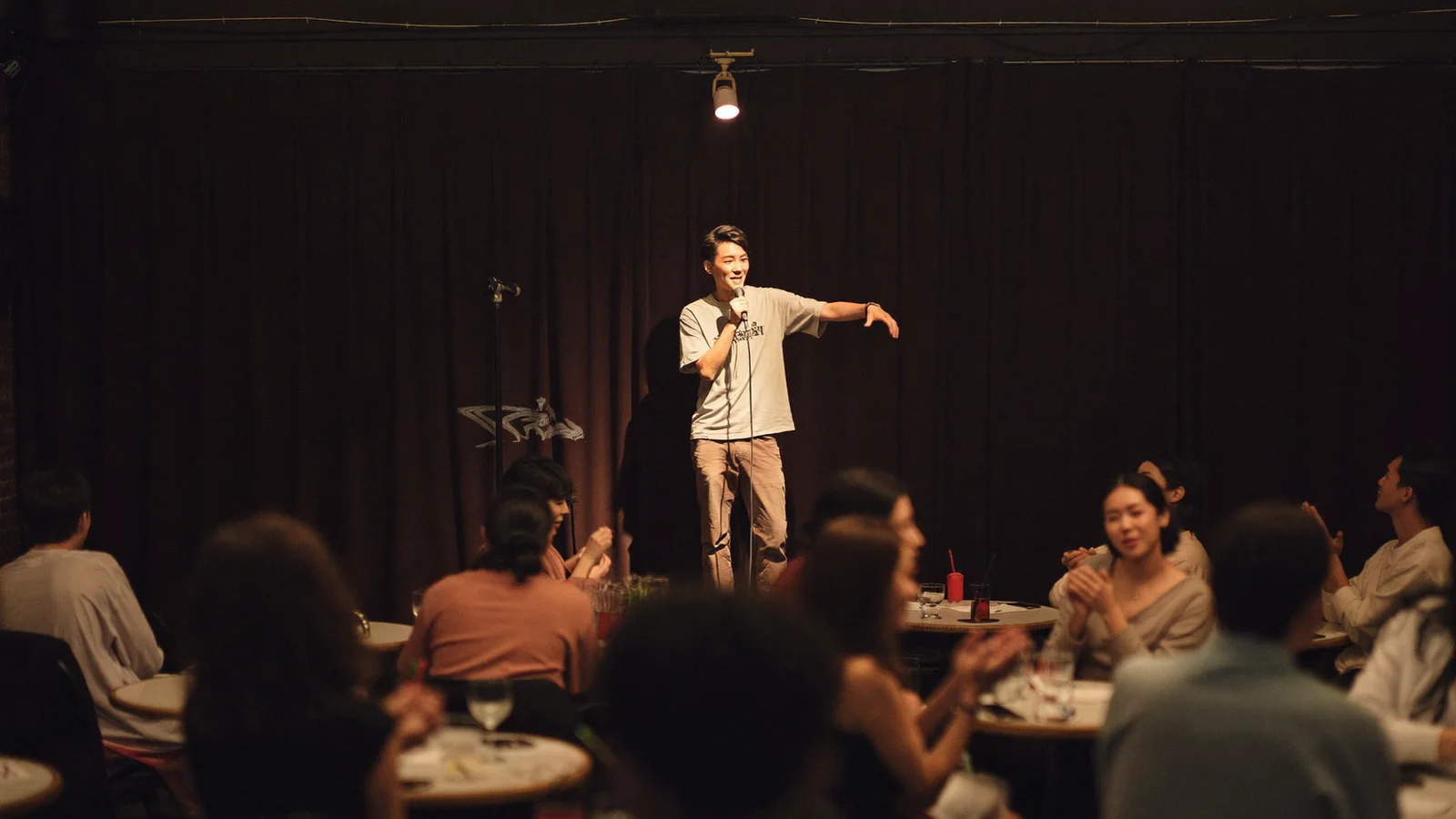 A moody comedy club stage with a single spotlight on a microphone stand and an intimate crowd of local Hong Kong Chinese patrons seated at small tables, softly blurred, illustrative image for this Hong Kong comedy show guide, not a photo of the actual venue or performer