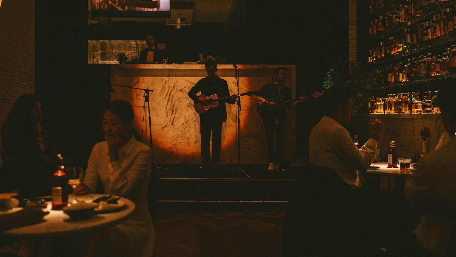 A dimly lit jazz bar interior with a small stage, warm amber lighting and cocktail glasses on a marble bar counter, local Hong Kong Chinese patrons seated at tables, illustrative image for this Take 5 H Queen's guide, not a photo of the actual venue or performers