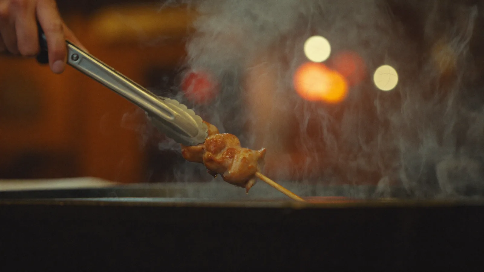 Skewers of grilled chicken yakitori sizzling over charcoal at a lively Japanese izakaya counter, with warm golden wood panelling and paper lanterns in the background and a local Hong Kong Chinese chef tending the grill, illustrative image for this Mong Kok yakitori restaurant guide, not a photo of the actual venue or dishes