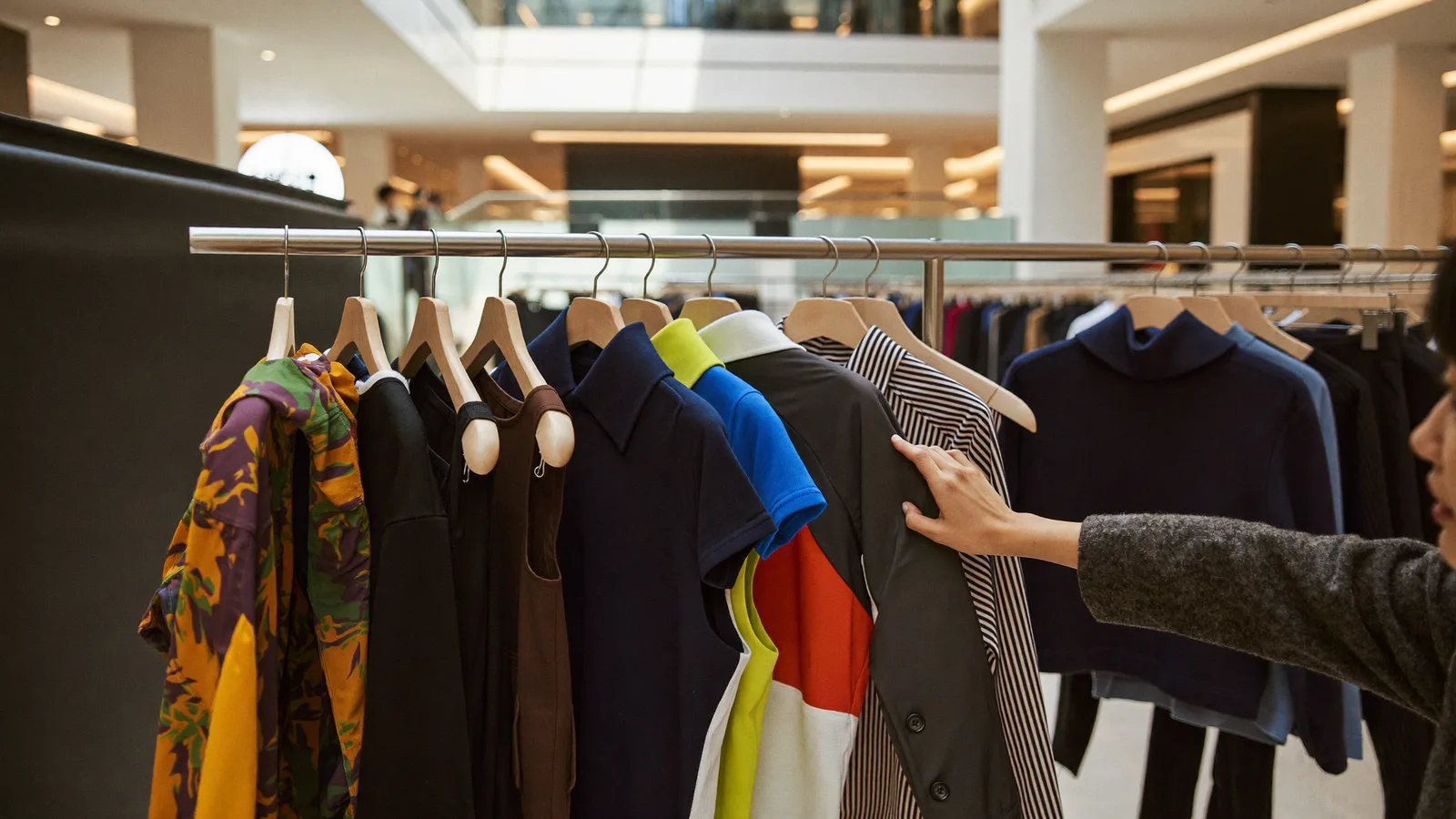 A stylish pop-up fashion retail display inside a bright Hong Kong shopping mall atrium, with clothing racks of contemporary streetwear and avant-garde designs, and a local Hong Kong Chinese shopper browsing and trying on a jacket, illustrative image for this Wear's HK fashion pop-up guide, not a photo of the actual event or designer collections