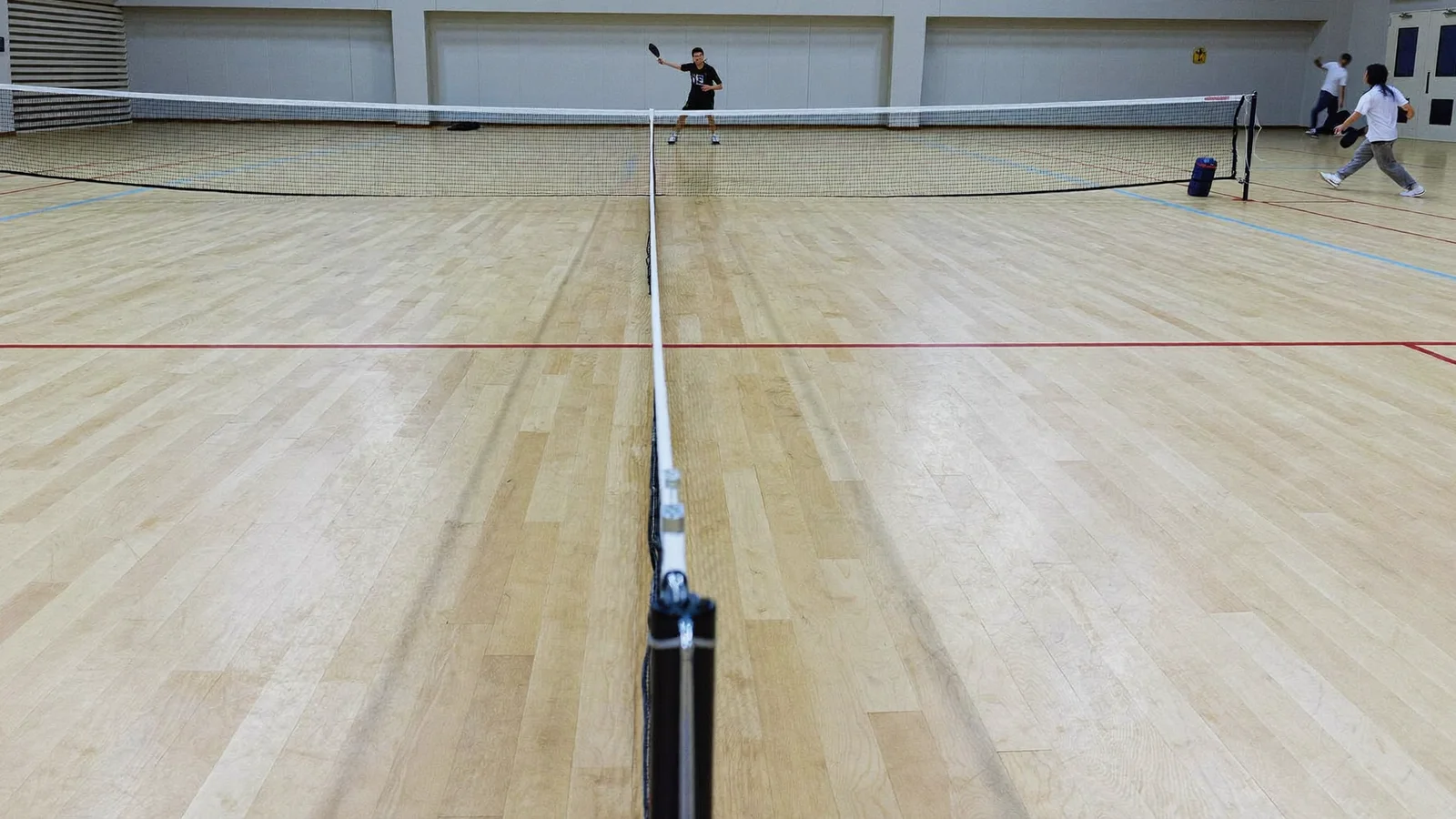 A pickleball paddle and ball resting on the net of an indoor court with painted court lines, local Hong Kong Chinese players rallying softly blurred in the background, illustrative image for this pickleball guide, not a photo of any specific venue