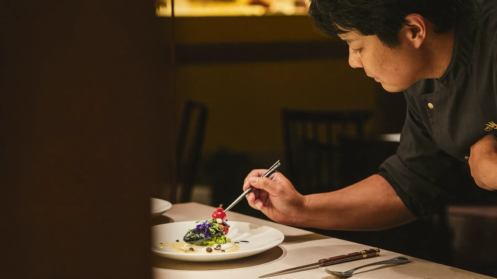 An illustrative editorial shot of an elegant Asian fine-dining tasting-menu course being plated by a local Hong Kong Chinese chef in a small, warm-lit restaurant, not a photo of the actual Whey x Upstairs Tasting Room dinner or its dishes