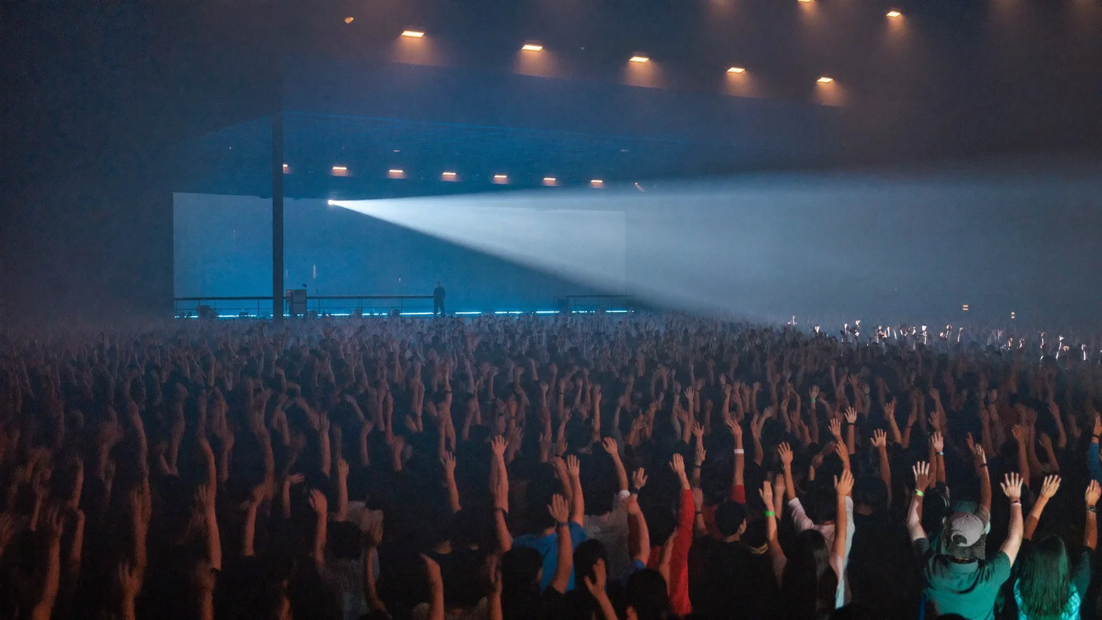 Illustrative image of a local Hong Kong Chinese concert crowd inside a large indoor arena, warm blue and amber stage lighting with a glowing stage in the background, no readable text or logos, not a photo of the actual show